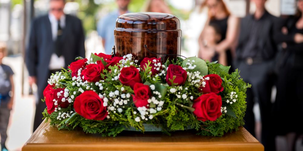 Urn displayed on a flower arrangement of red roses during a memorial service at a Funeral Home and Cremations in Milwaukee, WI, with grieving family in the background.